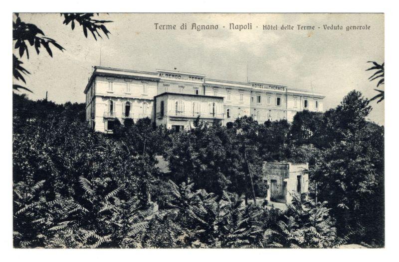 The image depicts a historical black and white postcard featuring the Hotel des Thermes in Agnano, Naples. The hotel is prominently situated on an elevated terrain surrounded by lush greenery, including ferns and dense bushes that suggest a botanical garden or park-like setting. Visible text at the top of the card reads "Terme di Agnano - Napoli - Hôtel delle Terme - Veduta generale," indicating the location in Italian as well as its English translation. The architectural style is classical with white facades, multiple stories, and numerous windows, some featuring shutters or balconies. A secondary building to the right has a simpler design compared to the main structure. An additional text "ALBERGO" can be seen on one of the buildings' roofs, likely indicating its function as an accommodation establishment. The overall scene conveys a serene atmosphere with natural elements surrounding the man-made structures, emphasizing tranquility and leisure associated with this historic spa town.