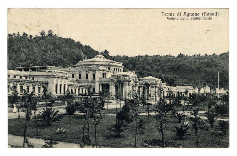 The image depicts a historic postcard showing the Termi di Agnano, also known as Napoli (Naples), which appears to be an old thermal bath and spa facility. It's captured in black and white with a sepia tone that suggests it is likely from early 20th-century Italy. The architecture of the building features neoclassical elements such as columns, arches, and a domed roof, indicating its grandeur.

The establishment sits amidst lush greenery including palm trees and well-manicured bushes, which adds to the tranquil atmosphere typical for thermal baths designed for relaxation and health treatments. A tram or trolley with open sides can be seen on tracks in front of the building, giving context to the era's mode of transportation as it is relatively early compared to modern electric trams.

In the background, a hill covered with dense vegetation provides a natural backdrop that contrasts with the structured elegance of the spa facility. The sky above appears overcast, and there are no pe