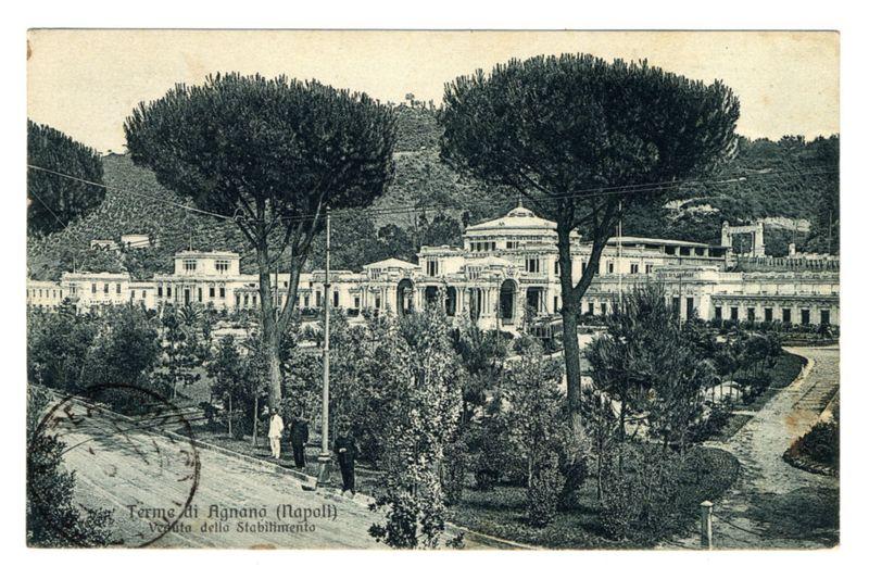 The image depicts a vintage postcard showing an old, grand building complex with classical architectural features such as columns and pediments. It is set in what appears to be early 20th-century attire, judging by the clothing of individuals walking on paths within manicured gardens or parks surrounding the buildings. The photo has a sepia tone typical for vintage photographs, giving it an aged look. Trees with dense foliage line up along the pathways and rise behind the structures, suggesting that this establishment is located in a hilly area. There are inscriptions at the bottom of the postcard which suggest some historical or geographical context to the location shown. The buildings seem stately and could be associated with luxury, such as possibly being a spa resort or an institution for recreation, given its name "Termes di Agnano" indicating thermal baths in Naples (Napoli).