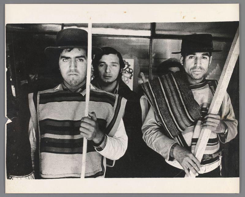 The image depicts three men standing side by side against a dark background. They are wearing hats, with two of them sporting wide-brimmed fedoras and one donning what appears to be a trilby or similar style hat. The man on the left is holding onto white vertical bars that suggest they might be in some sort of barred structure, possibly handcuffs around their wrists.

The men's attire varies: the first two have horizontal striped vests over dark shirts, while the third wears an ornate vest with a mix of stripes and patterns, along with what seems to be a scarf or shawl draped across his shoulders. The man in the middle is holding onto one bar with both hands near his chest level, suggesting he might not be free from some form of restraint.

The expressions on their faces are serious; none are smiling, which adds an element of tension or gravity to the scene. Their eyes are focused and directed towards something out of frame, possibly capturing a moment where they're aware of being 