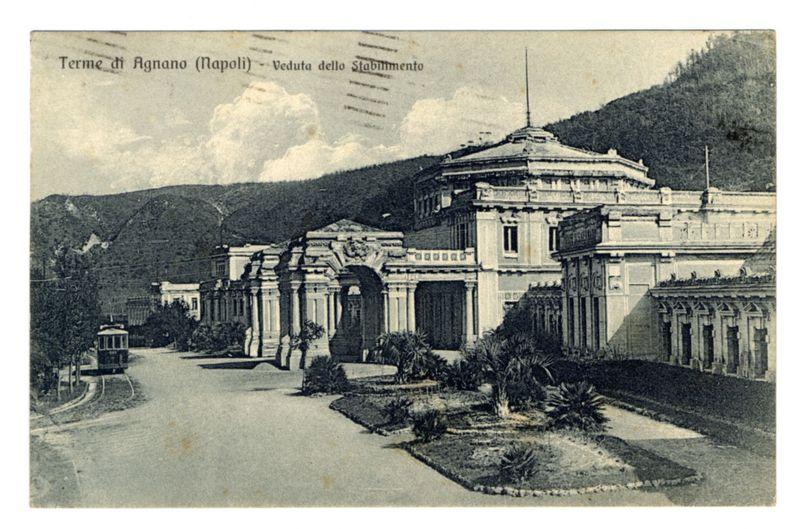 The image depicts a vintage postcard showing an architectural structure labeled "Terme di Agnano (Napoli) - Veduta dello Stabimento." The building is grandiose with classical architecture elements, including tall columns and intricate designs. It appears to be situated in the hills of Agnano near Naples as indicated by the text on the image.

The foreground features a street leading up to the main entrance where a tram or trolley car is visible at an intersection, suggesting it was taken during a time when such public transportation systems were common. The surrounding area includes neatly maintained gardens with pathways and various plants that indicate landscaping for aesthetic appeal. In the background are hills covered in greenery, possibly indicating the location's proximity to natural landscape.

The image conveys historical significance as postcards of this nature often served to promote tourism or commemorate notable places during their time period. The absence of modern el