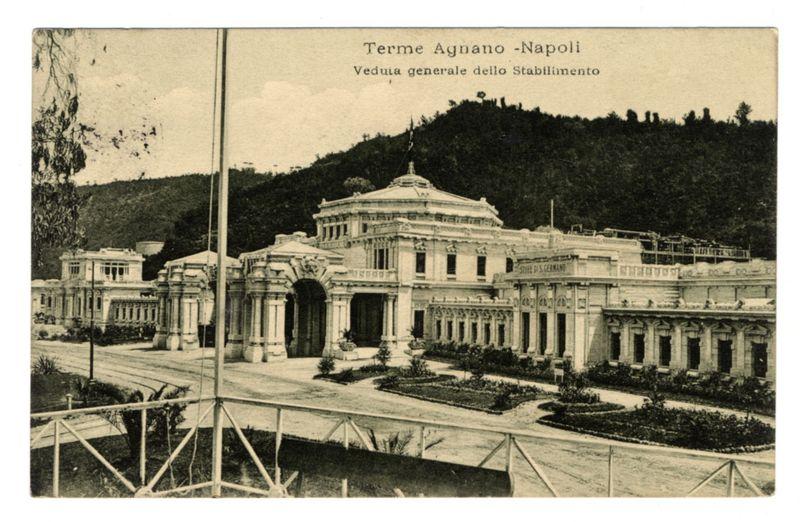 The image displays a sepia-toned postcard featuring an old, grand building with classical architectural elements such as columns and pediments. The structure is labeled "Terme Agnano - Napoli" indicating it's located in Naples at the Terme Agnano Thermal Baths. It appears to be viewed from a distance given the perspective that captures both the main building and several wings extending on either side, set against a backdrop of hilly terrain with sparse vegetation. There are some trees or bushes visible near the foreground, and what looks like fencing partially obscures parts of the scene in the lower part of the image. The sky is overcast, suggesting it might be an older photograph capturing historical architecture rather than current tourist attractions. No people can be seen, which could indicate this photo was taken when fewer visitors or during off-peak hours for such a public venue.