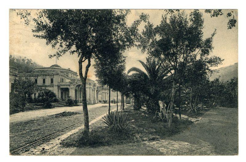 This image depicts a vintage black and white photograph of an old building with classical architectural features, such as columns and ornate detailing. The structure is surrounded by lush greenery including palm trees, shrubs, and large-leafed plants that suggest a tropical or subtropical climate. A well-maintained pathway runs through the garden area leading towards the building entrance. To the left of the photograph, there appears to be railway tracks intersecting with the path, indicating transportation access during its time. The background shows rolling hills or mountains in the distance under an overcast sky. There are no visible human figures within this image; it focuses solely on the architecture and natural environment.