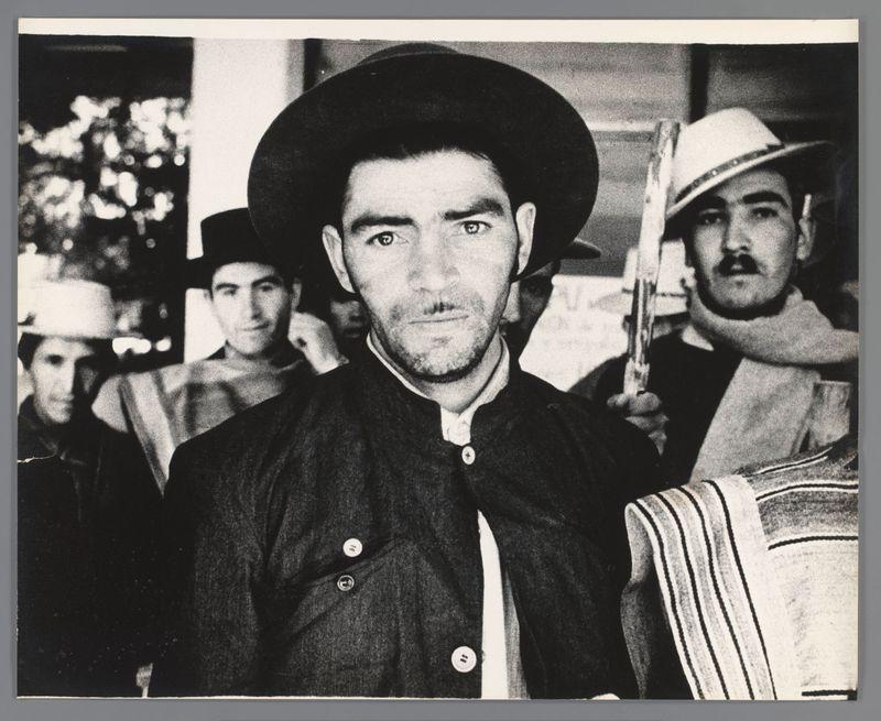 The image is a black and white photograph featuring several individuals who appear to be involved in some sort of organized activity, possibly related to labor or protest. The main subject is positioned centrally and dressed in traditional attire with a dark jacket adorned with buttons and wearing a hat that covers the head completely. This individual has a serious expression on their face.

Behind this central figure are others; one person is partially visible at the top right corner of the image, holding what looks like a firearm or a tool pointed towards something out-of-frame to his left side. Another man in traditional clothing with a hat and mustache stands behind him, also looking directly into the camera but less intensely than the central figure.

The background shows blurred figures that seem engaged in their own activities, possibly observing the event from distance. The overall composition suggests this photograph was taken at an outdoor gathering or protest during dayl