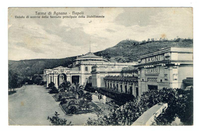The image depicts a historical postcard showing the "Terme di Agnano" in Naples, an Italian thermal spa facility. The photograph is monochrome and displays classical architecture with grand columns, elaborate facades, and manicured gardens. It appears to be from several decades ago based on its style of presentation. On the right side of the image, a building reads "BAGNI 2° CLASSE," indicating it was likely classified as a public bathhouse or spa during that period. The landscape in the background suggests the location is elevated with vegetation and hills visible further back. There are people walking along what seems to be an approach road leading up to these buildings, providing scale and context for the size of this establishment.