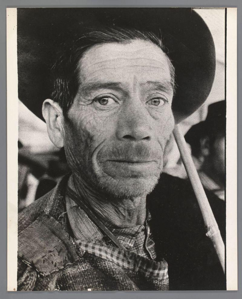 The image is a black and white portrait of an older man with weathered skin, indicative of significant outdoor labor or exposure. He wears a wide-brimmed hat that casts shadows on his face, enhancing the lines around his eyes which suggest age and experience. His gaze directly meets the viewer's eye, conveying intensity and perhaps a sense of defiance against the elements he has likely faced throughout life.

He is dressed in what appears to be rugged work attire, with layers suggesting practicality over style—perhaps indicative of manual labor or agricultural employment. The checkered pattern on his collar hints at a uniform worn by workers from that era.

The background features blurred figures and objects which give the impression of an outdoor setting, possibly during daylight due to the brightness around him despite the monochromatic nature of the photo. His expression is somber yet poignant, capturing not only physical characteristics but also hinting at life's hardships thro