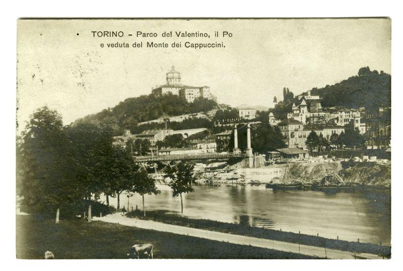 The image depicts an old postcard showing a historical scene of Turin's Parco del Valentino, with the Po river and the view from Monte dei Cappuccini. The card features black-and-white photography, capturing European architecture along the riverside, including buildings on terraced hillsides that rise above the water. Prominent structures include what appears to be a tower or lookout building atop one hillside in the background.

Trees line the riverbank and a solitary cow grazes near the water's edge at the forefront of the image. The lighting suggests it is daytime with clear skies, as shadows are visible on trees and ground features.

There is text overlaid on the upper portion of the card which translates to "Turin - Parco del Valentino, Po River View from Monte dei Cappuccini." This indicates that the postcard was likely distributed in a period when photography had become accessible for personal mementos. The overall tone and condition suggest an antique item with historical s