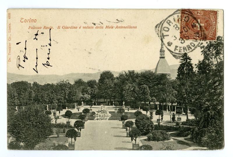 The image depicts a vintage postcard showcasing the gardens of Palazzo Reale in Turin, Italy. The card features an old photograph with black and white tones capturing meticulously maintained garden paths leading to fountains surrounded by trees and shrubs. In the background stands a prominent dome-shaped structure, likely part of the Mole Antonelliana museum. Handwritten text on the left side appears to be inscriptions or signatures in cursive script. The top right corner displays an ornate red stamp with "TORINO" printed prominently across it, indicating Turin's name. A postage stamp is affixed at the upper edge, bearing a denomination of 2 lire and adorned with intricate designs including what seems like coat-of-arms imagery. Below this seal, additional text in Italian mentions Palazzo Reale and highlights that both the garden and Mole Antonelliana can be seen from it.

The card's left side has printed details such as "Dr. Trinkler Co., Lipsia" indicating a printer or distributor b