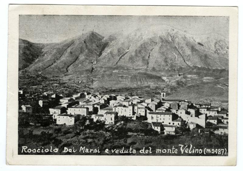The image depicts a black and white photograph of a small village nestled in the foothills with mountains looming behind. The buildings appear to be traditional, likely made from stone or plastered mud bricks, exhibiting simple rectangular shapes with pitched roofs. A church tower is visible on one side, suggesting religious significance within this community.

Inscription at the bottom reads "Rosciolo Dei Marzi e Veduta del Monte Velino (m.2487)," indicating that it's a historical document from Rosciolo dei Marsi and a view of Mount Velino taken around 1936, measuring approximately one meter in height.

The terrain is rugged with sparse vegetation, hinting at the arid climate or high-altitude setting typical for such Italian landscapes. The overall quality suggests that it might be an old photograph captured using early photographic techniques, likely from the late nineteenth century to mid-twentieth century based on its appearance and the caption provided.
