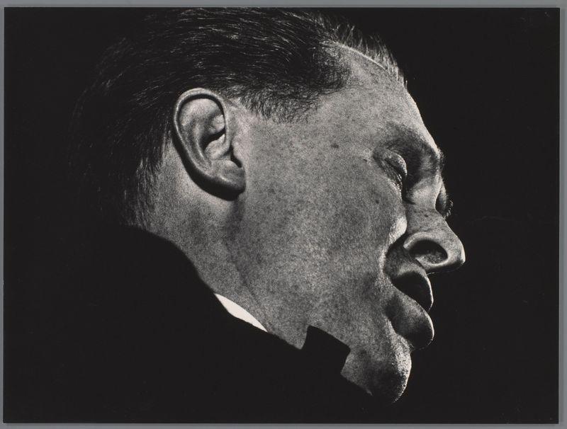 The image is a black and white photograph of the profile view of an individual. The person appears to be wearing glasses, which are reflected on their face in such a way that suggests intense focus or concentration. Their ear and hairline can also be seen clearly due to this reflection.

This particular photo seems like it could have been taken during a musical performance as suggested by additional information provided: "Herb Ellis op het concert Jazz at the Philharmonic, Scheveningen" (Ed van der Elsken) from 1958. This indicates that Herb Ellis is likely performing or perhaps even leading this session.

The background of the image has been completely darkened to highlight the subject's profile, creating a dramatic and focused composition where all attention is drawn towards his face and expression which conveys deep engagement with what he might be doing at that moment - singing, speaking, playing an instrument.