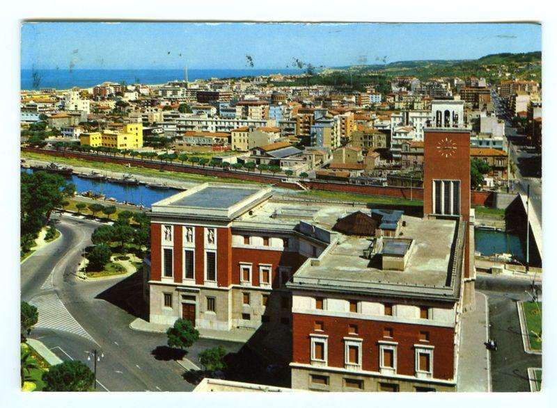 The image presents an expansive view of the cityscape, capturing a blend of architectural styles indicative of urban development. In focus is a distinctive red brick building with white trim and large windows on its facade, suggesting it could be institutional or governmental in nature. This structure has a prominent clock tower to one side, reinforcing its significance within this setting.

Adjacent are modern buildings featuring clean lines and glass facades that reflect the surrounding environment, blending contemporary design into an older urban fabric. The presence of greenery—trees lining streets—is noticeable but limited due to the predominance of built-up areas.

A significant feature is a river or canal running through the scene with boats moored along its banks, hinting at local commerce and possibly recreational activities. Bridges span across this waterway, connecting different parts of the city that are densely packed together in colorful buildings—ranging from yellow 