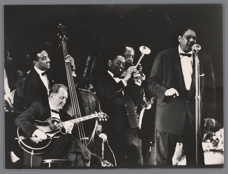 The image captures a moment from what appears to be a live jazz performance, featuring five musicians in formal attire. In the foreground on the left, an individual is playing a guitar with his eyes closed and leaning into the instrument, suggesting deep engagement or emotion during play. To the right of this guitarist, another musician holds a trumpet near his mouth, possibly having just played or preparing to play. Behind him stands a bass player in mid-motion, indicating active participation. Further back on stage is an individual playing what seems to be a double bass with concentration and perhaps leading others harmonically through their piece.

The central figure of the image is at the microphone stand singing passionately into it; his facial expression conveys intensity typical of jazz vocal performance. His hand gestures add dynamism, implying interaction or emphasis in his song delivery. The background consists mainly of dark shades which draw focus to the performers and am
