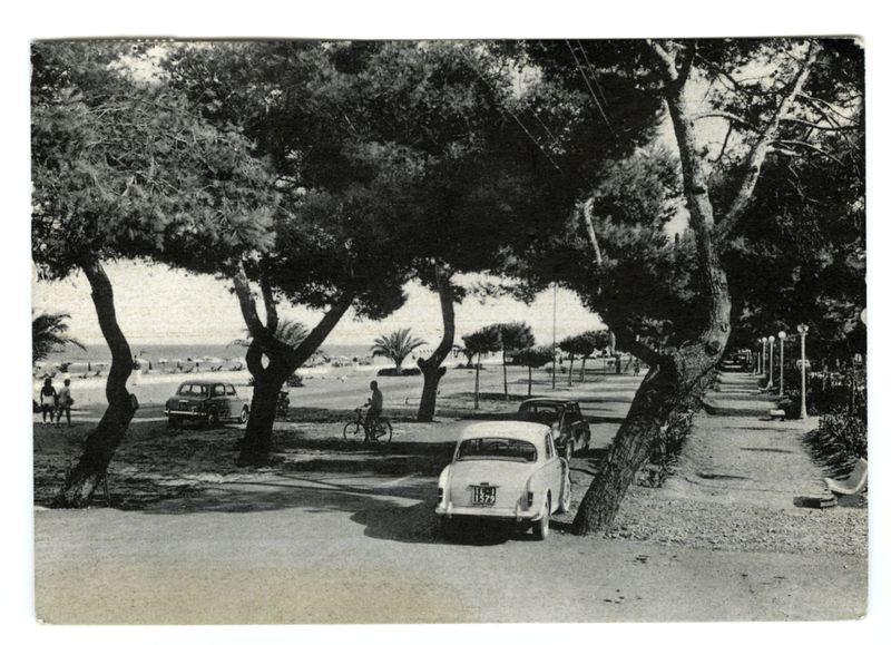 The image depicts a vintage black and white photograph of a coastal scene, likely from the mid-20th century. The main subject is an open area with a beach in view on the left side where people are scattered along the shore, some walking or standing near the water's edge. A prominent feature is the expansive body of water that stretches to the horizon under a clear sky.

In the foreground, there is a park-like setting with trees casting shadows over what appears to be an asphalt path lined with lamp posts and benches for relaxation. Several vintage cars are parked on this road; one white Lancia car in particular stands out due to its size relative to other vehicles and visible registration plate. The overall atmosphere suggests leisurely activity, possibly a public park near the sea.

The photograph's monochromatic tones convey an aged quality, with varying shades of gray that suggest depth and contrast within the image. There is no direct indication of specific individuals or perso
