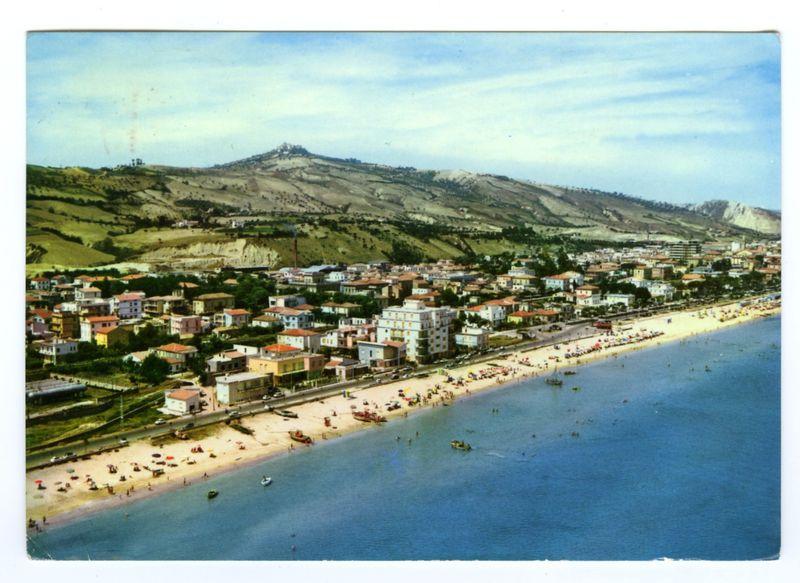 The image depicts an aerial view of a coastal town, featuring a beachfront with numerous people engaging in various activities. The sandy shore is dotted with umbrellas and sun loungers, indicating recreational use by visitors or residents alike. A row of multi-story buildings lines the coastline, exhibiting different colors such as white, beige, red-roofed structures, which suggest residential or commercial establishments catering to tourists.

Beyond the town, a series of rolling hills ascends into what appears to be a hilly countryside landscape with sparse vegetation and some cultivated land in patches. The backdrop reveals a prominent hilltop structure that could potentially serve as an observation point or landmark for the area. Above it all is a sky filled with soft clouds, hinting at fair weather conditions conducive to outdoor activities.

The clarity of the photo suggests that it was taken from a considerable height above ground level, likely by helicopter, which also cor