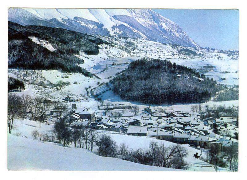 The image depicts a snowy landscape with a small village nestled in the hills. The scene is dominated by snow-covered slopes and trees, as well as rooftops of buildings dusted lightly with white. A prominent feature in this area appears to be an elevated rocky outcrop amidst the lower terrain covered with snow.

In the background, towering mountains are visible, their peaks blanketed with snow suggesting a cold climate or winter season at least during which time the photograph was taken. The sky is clear and blue, indicating fair weather conditions above such harsh landscapes below.

This photo captures not only a picturesque rural scene but also showcases how human settlements can coexist harmoniously in challenging natural environments like this one characterized by mountainous terrains covered with snow.