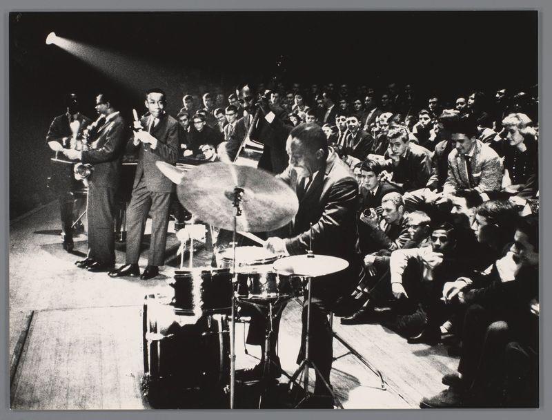 The image captures a dynamic moment during a jazz performance with multiple musicians on stage. A central figure is energetically striking the snare drum, highlighted by dramatic spotlighting that accentuates his action and the movement of his limbs. Surrounding him are other members of what appears to be an ensemble; some hold brass instruments such as trombones or trumpets, while others play percussion like a bass drum and cymbals. The audience is in various states of engagement—some leaning forward attentively with hands near their mouths suggesting applause or anticipation, others sitting back more passively observing the performance. The image exudes an atmosphere of intense live music performance from what could be mid-20th century judging by attire and photographic style; it's black-and-white film that captures a significant cultural event possibly in Amsterdam during 1958 as per provided context.