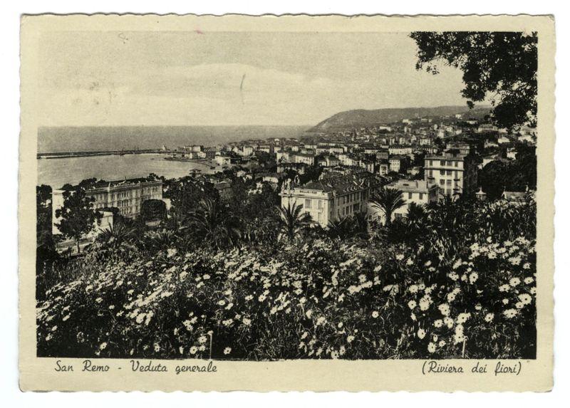 The image is a sepia-toned photograph of the town San Remo, as viewed from an elevated perspective. It captures various buildings with European architectural styles, including multi-story structures with flat roofs and terracotta tiles, typical for Mediterranean regions like Liguria in Italy where San Remo is located. The skyline features lush greenery, palm trees, and dense foliage at the lower part of the image that gradually gives way to urban development as it ascends toward more residential areas.

In the background, a calm sea or bay can be glimpsed with some structures along its edge suggesting port facilities. A hill rises in the far distance beyond the town's skyline, adding depth to the composition and hinting at further geographical features outside of the captured view. The photograph has text on it indicating that this might be part of an old postcard series or a historical documentation.

The bottom left corner contains handwritten script labeling "San Remo - Veduta g
