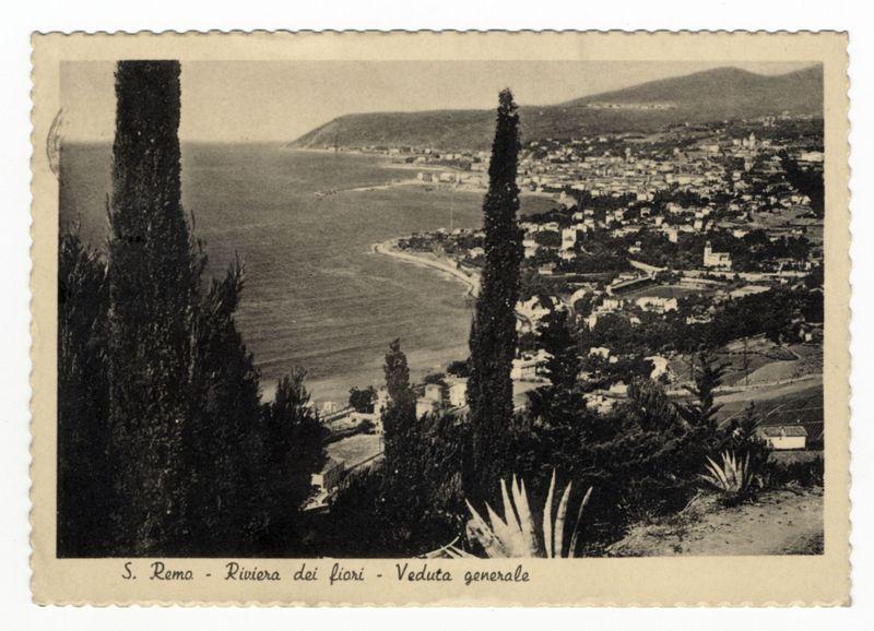 The image is a sepia-toned, vintage photograph that appears to be from the early 20th century. It depicts an aerial view of the coastal city of Sanremo (S. Remo), located on the Riviera dei Fiori in Italy. The perspective shows the town extending along the shoreline with clusters of buildings and a clear separation between urban areas and more open spaces, possibly agricultural land or parks.

In the foreground, two large cypress trees are prominently featured on either side, framing the view towards the sea. Behind them is an agave plant, adding to the Mediterranean flora ambiance. The coastline curves gently along what seems like a peaceful beach area with waves lapping against the shore visible in the distance.

The top of the photograph has text that reads "S. Remo - Riviera dei fiori - Veduta generale," which translates from Italian as "Sanremo - Flower Coast - General View." The image appears to be a postcard or a similar printed material, given its border and textured appear
