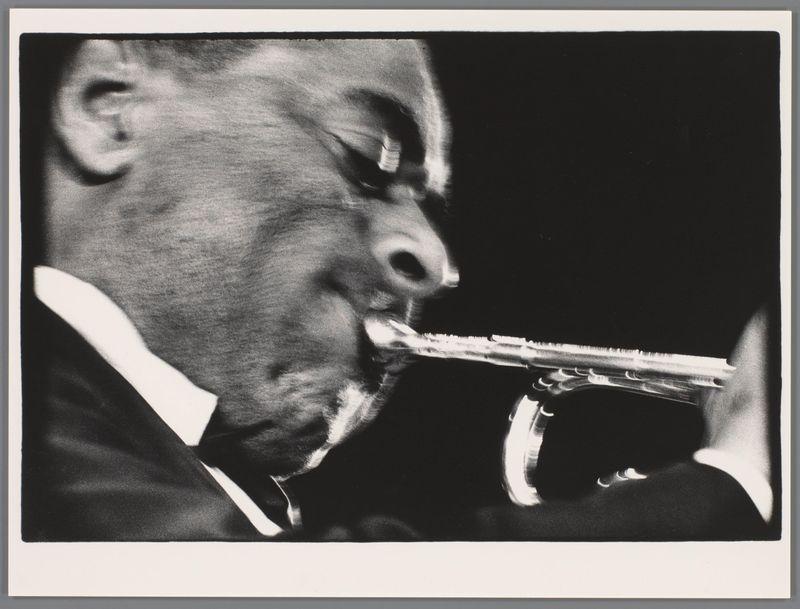 The image is a black and white photograph of an individual playing the trumpet. The focus is on capturing the intense expression and motion as they play, with light accentuating their face and mouthpiece while casting deep shadows in other areas. This style suggests it might be from the mid-20th century or created to emulate that era's photography aesthetics. There are no discernible background elements due to a tight framing of the subject; all attention is drawn toward the musician’s performance.