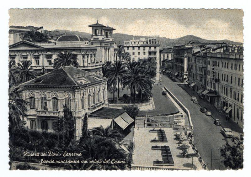 The image is a black and white photograph depicting an urban scene with classical architecture. It shows several buildings, including what appears to be the "Riviera dei Fiori" in Sanremo as indicated by text at the bottom of the photo. The prominent structure has multiple levels, symmetrical windows, and decorative cornices on its facade. Palm trees line a street that extends into the distance where various other period-style buildings flank it.

There are also what seem to be small gardens or courtyards with potted plants near one building in the foreground. A few cars from an earlier era parked along the side of the road, suggesting this photo is historical. The architecture features balconies and large windows typical for European urban environments at that time.

The top left corner has a torn paper edge effect indicating it may be part of a postcard or old photograph with some wear over time.
