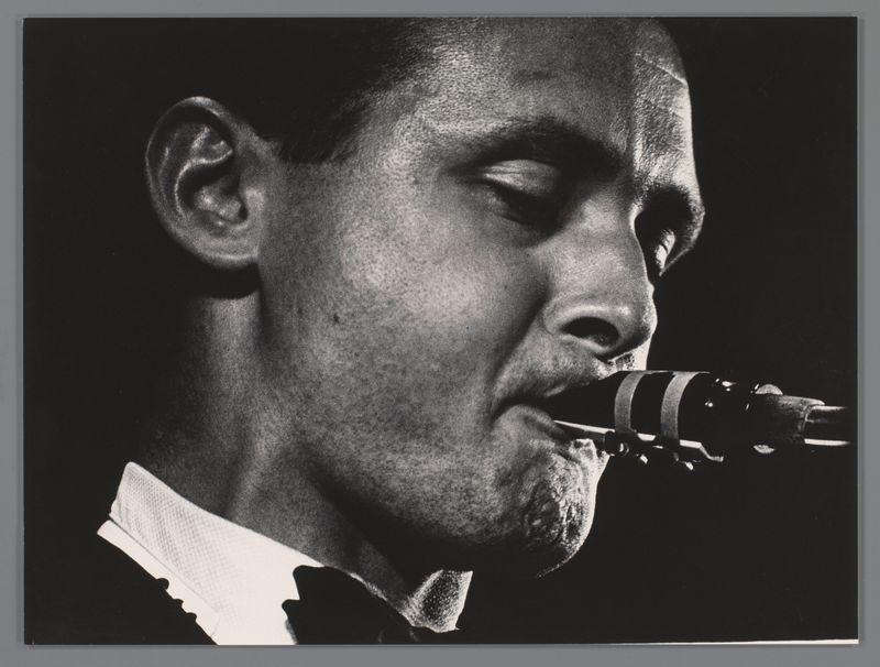 The image is a black and white photograph depicting an individual playing the saxophone. The focus is on capturing the musician's intense expression while performing, with close-up shots of their face, showcasing details like wrinkles around the eyes suggesting concentration or emotion in his performance.

In this particular snapshot from Ed van der Elsken’s 1958 photo series "Stan Getz op het concert Jazz at the Philharmonic" taken in Scheveningen, it captures a moment that likely represents Stan Getz's improvisational skills and emotional depth. This image is part of a larger collection showcasing jazz performances during this iconic event.

The saxophone itself appears prominently against the darker background, emphasizing its importance as an instrument within this genre. The lighting accentuates certain features such as the musician’s facial contours and highlights textures like skin folds and fabric creases in his clothing, adding to the overall moodiness of the scene.
Stan 