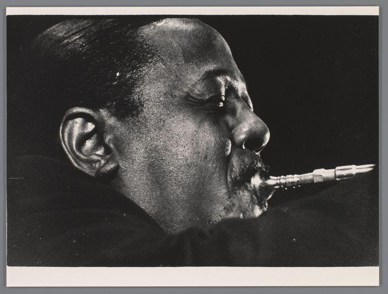 The image is a black and white photograph capturing an individual in the midst of playing a saxophone. The focus is on the upper body, showing detailed features such as skin texture, facial contours, and fingering technique on the instrument. There's no discernible background; it appears to be taken against a neutral or dark backdrop which directs attention solely towards the subject and their activity.

This image can serve as an evocative portrayal of jazz musicianship from the mid-20th century, potentially reflecting Roy Eldridge during his performance at "Jazz at the Philharmonic" in Scheveningen, captured by Ed van der Elsken. The historical context provides insights into the cultural and artistic environment of that time within the realm of American Jazz music.

Without any explicit textual information or identifying markers present directly on the image itself, it relies heavily on indirect cues to infer its narrative and significance.