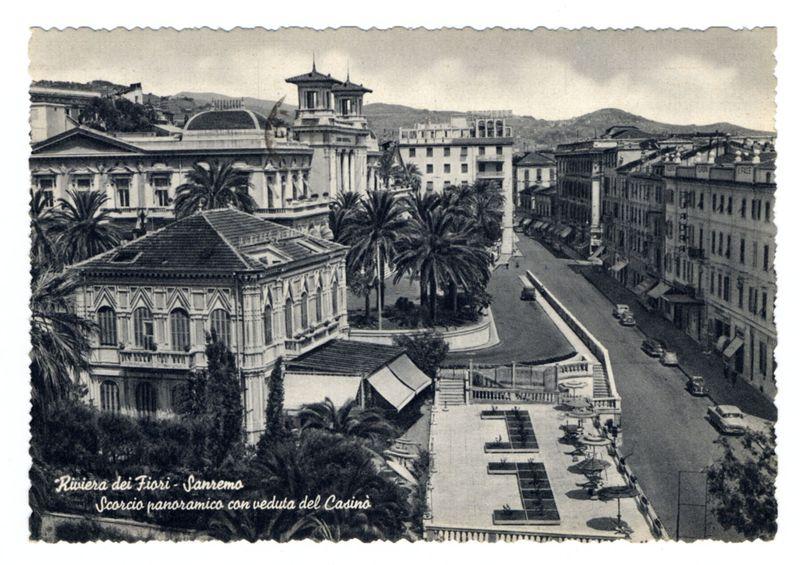 The image is a black and white photograph of Riviera dei Fiori in Sanremo, Italy. It appears to be an old postcard featuring European-style architecture with ornate facades, large windows, and decorative details such as balconies and cornices. The buildings have classical influences evident through their symmetry, tall columns, and grandeur.

In the foreground, there's a paved area with rectangular planters housing plants or small trees arranged in rows, possibly part of an urban park or plaza. A wide street runs parallel to this square, lined with parked cars indicative of mid-20th-century transportation style. The buildings along both sides have multiple floors and display commercial signage, suggesting active businesses on the ground floor.

Palm trees dot the landscape near a prominent building at one end, which stands out due to its height and classical design elements including columns and towers that may signify an institutional or historical significance. In the background,