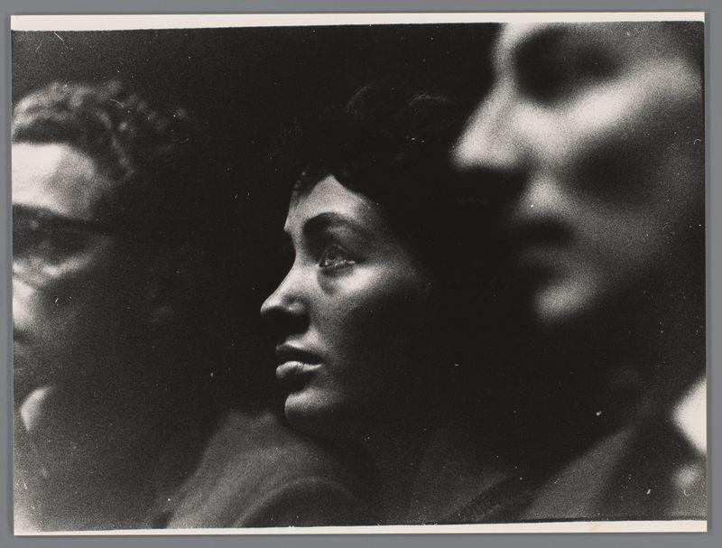 The image is a black and white photograph capturing a moment at what appears to be an event or concert. In the foreground, there's a close-up of individuals with their faces partially illuminated by stark lighting against a dark background, emphasizing dramatic shadows that obscure most facial features. This creates a strong contrast between light and shadow which adds intensity and depth.

In the mid-ground, two people are standing in profile facing away from the camera towards an event or audience out-of-frame. Their heads and upper shoulders are visible with subtle details of their profiles discernible despite being predominantly engulfed by shadows. The person on the right is more prominently featured than the one to the left due to better exposure.

The background suggests a crowded setting, likely inside given the close proximity of individuals that can be seen at various levels within what appears to be an elevated platform or tier seating arrangement typical for concerts or