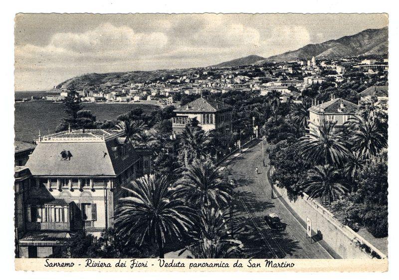 The image depicts a black and white photograph of the coastal cityscape known as Sanremo, located on the Italian Riviera. This postcard shows an aerial perspective overlooking the "Riviera dei Fiori" or Flower Coast area near Portofino Bay with palm trees lining what seems to be via San Martino street below. The architecture includes European style buildings with a prominent one in the foreground featuring decorative windows and balconies, indicative of early 20th-century design aesthetics.

In the background, hills rise behind residential structures leading towards more urbanized areas filled with various types of buildings ranging from houses to possibly commercial or public facilities. The sea is visible on the left side, showing a calm expanse of water extending into the horizon under an overcast sky that suggests either early morning light or late afternoon.

The postcard bears text in Italian and French which translates as "Sanremo - Riviera dei Fiori - Panoramic view from Sa