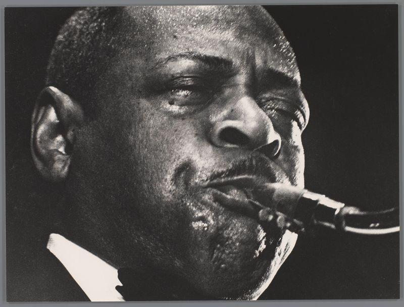 The image is a black and white photograph capturing the intense expression of an individual playing jazz saxophone. The musician's face fills most of the frame, with fine details such as wrinkles around his eyes and mouth accentuating the emotional intensity of the performance. His cheeks are puffed out as he blows into the saxophone, which appears in sharp contrast against a dark background. A bright highlight is cast across part of the saxophone near where it meets the player's lips, drawing attention to this action. The lighting creates deep shadows and highlights on his face, adding depth and texture that emphasize the contours of his features. This monochromatic image focuses solely on the performer, with no distractions in the background or surrounding environment visible.