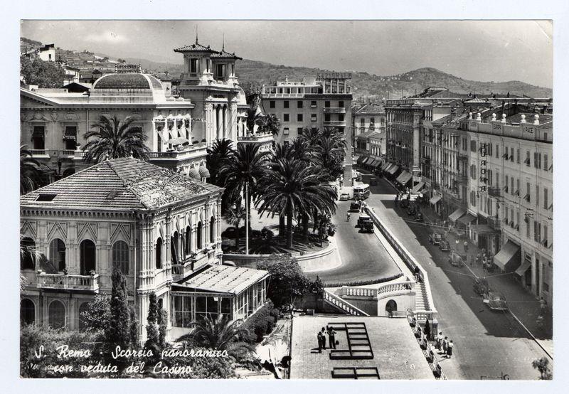 The image is a black and white historical photograph showing an urban street scene. It appears to be taken from an elevated perspective, overlooking a road lined with buildings on both sides. Prominent features include palm trees planted along the median strip of the roadway, which suggests warm climate typical of Mediterranean regions.

The architecture varies but includes classical elements like columns and domes, as well as modernist styles such as Art Deco seen in tall commercial buildings adorned with signage. The presence of people walking on sidewalks indicates a bustling urban environment.

A bridge or underpass is visible to the right side where multiple pedestrians are crossing from one street level to another. At least two vintage vehicles can be spotted driving down the road, suggesting that this photo was taken several decades ago when such models were common in public use.

The text at the bottom of the image reads "S. Remo - Scario panoramico con veduta del Casino,