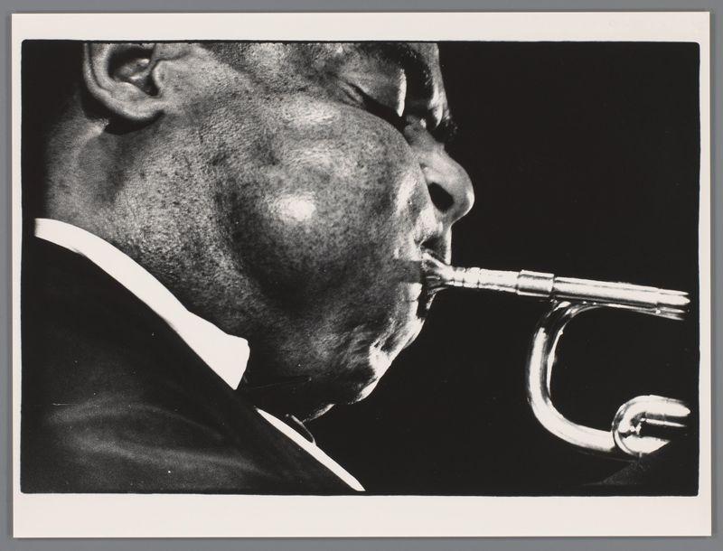 The image captures a close-up of an individual playing the trumpet. The focus is on the musician's face, particularly around his cheeks and lips as they form into a rounded shape to produce sound through the instrument. We see only part of the person’s ear on one side and hairline visible above the temple area. His eyes are closed or nearly shut, suggesting deep concentration or immersion in performance. The trumpet is prominently displayed against a dark backdrop that accentuates its shiny brass surface with smooth curves and reflective gleam indicating it's well-maintained or new.

The black-and-white tonality of this photograph adds to the classic feel, possibly hinting at an older time period associated with jazz music when such monochrome imagery was prevalent. The absence of color directs attention solely on texture contrasts—the roughness of skin versus the smooth trumpet—and emphasizes expressions and emotions conveyed through body language or instruments.

There is a stark