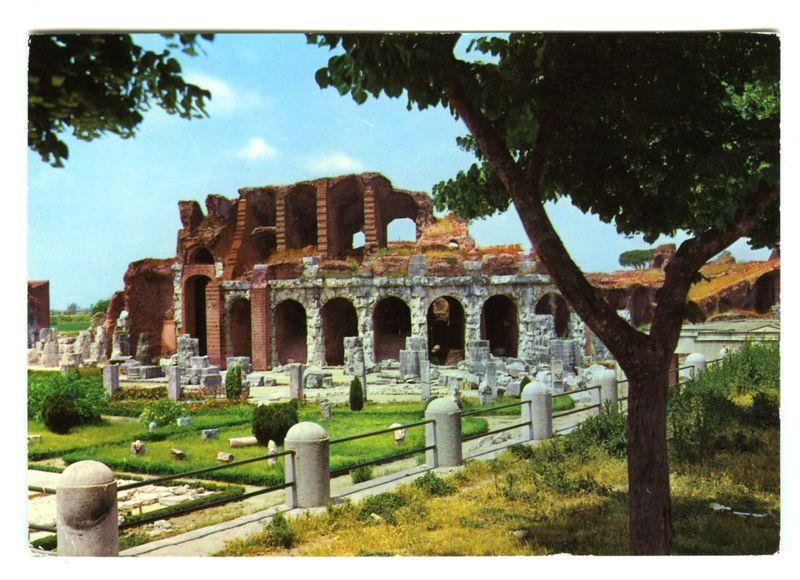 This image features a historical Roman amphitheater ruin under bright daylight. The structure is composed of large, weathered stone blocks in hues of red and gray, indicative of ancient construction materials like volcanic tuff from the nearby Mount Vesta. Arched openings line one side, suggesting where seating or decorative elements might have been located. Surrounding grassy areas with manicured hedges provide a contrast to the earth-toned stonework, highlighting pathways that lead through remnants of architectural columns and fragments.

The photo is taken at an angle from within what appears to be another historical site, possibly indicative of Roman gardens or a similar space, as suggested by garden-like structures in the foreground. The scene includes green foliage partially obscuring the view on one side, adding to the ambiance of preserved antiquity amidst nature's reclamation.

The overall setting conveys an atmosphere of serene preservation with visible signs of time and 