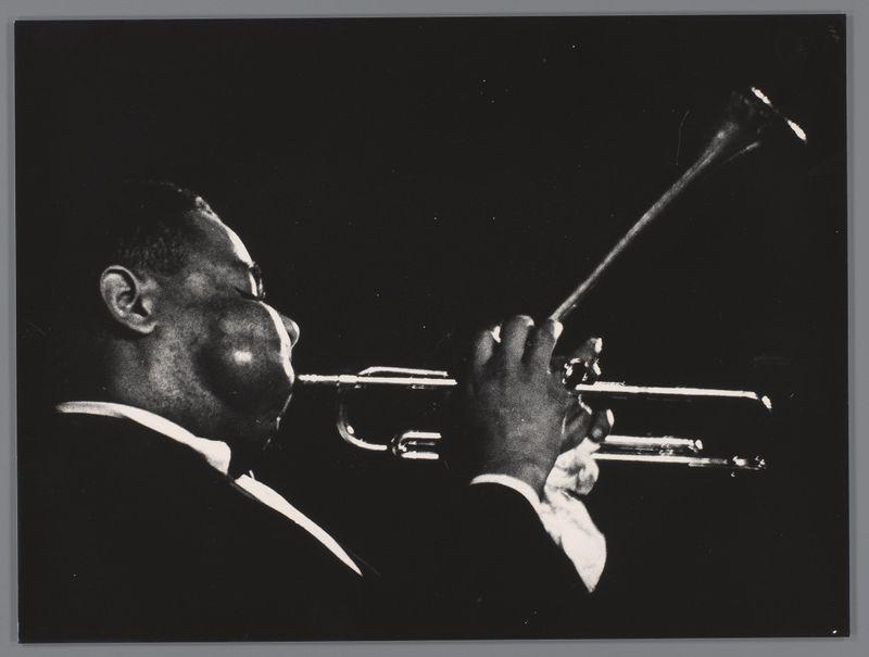 This image is a black and white photograph capturing an individual playing the trumpet. The musician, possibly male given their physique, appears to be deeply engrossed in performance, with his head bowed slightly towards the instrument as he blows into it. His right hand holds what seems to be a mute near the valves of the horn while his left fingers are positioned on the valve keys. He is dressed formally, likely wearing a dark jacket and white shirt which suggests an indoor concert setting or formal event. The background is completely black, providing high contrast that highlights the trumpet player's silhouette against it. There is no visible audience in this shot; thus, we can't comment on their reaction to his performance.

The image has a dramatic quality due to its monochromatic palette and strong chiaroscuro effects which emphasize textures like the musician’s skin tones and the reflective surface of the brass instrument. The composition centers around the trumpet player's h