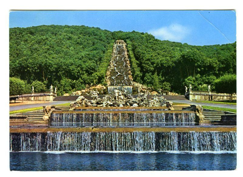 The image depicts a grand fountain set within an expansive landscaped area. The centerpiece is a tiered cascade of waterfalls, with multiple levels descending from the top to lower pools below. Atop this structure sits a sculptural ensemble featuring several large figures and animals in various poses, possibly depicting mythological scenes or historical events. These sculptures appear aged but are still detailed enough to be distinguished.

The fountain is surrounded by classical architecture elements such as balustrades with statues of draped figures at the ends, which suggest an elegant design typical of European palaces or estates. The backdrop consists of a lush green hillside that rises steeply behind the waterfall, enhancing the sense of elevation and grandeur for the cascading water.

A wide expanse of calm blue-green waters lies in front of the main fountain structure, reflecting some parts of it. On either side of this central feature are parallel lines of steps or balustr