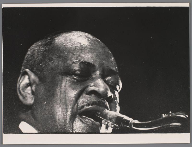 The image is a black and white photograph capturing an intense moment of performance. It depicts the close-up profile of a tenor saxophonist, likely Coleman Hawkins given the context provided in the alt text. The musician's face shows deep concentration as he plays into his instrument with fingers on the keys, blowing air through it. His expression is one of immersion and passion for music. There are subtle light reflections highlighting parts of his facial features such as his nose and lips, which add a sense of depth to the image.

The background is dark and lacks any distinguishable details, ensuring that all focus remains on the saxophonist's performance. The monochrome palette emphasizes textures, especially in the musician’s skin, creating a dramatic effect appropriate for conveying emotion through music. No other objects or people are visible in this tightly framed shot; it focuses solely on the artist and his instrument.

The photograph carries an artistic quality with its 