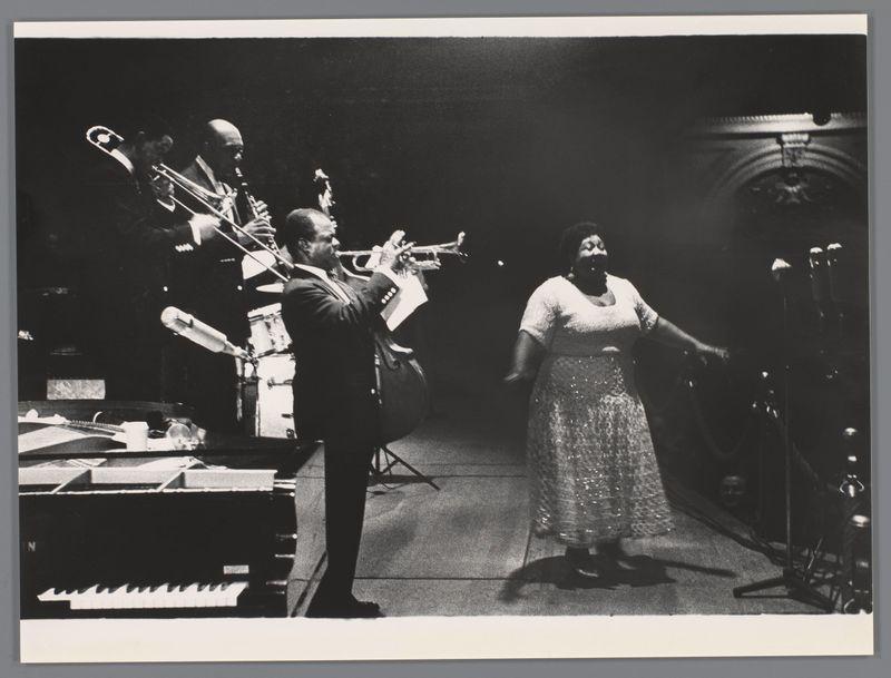 A black and white photograph depicting a musical performance, likely in an intimate setting such as a jazz club. In the foreground, two musicians are captured mid-performance: one playing trumpet with intense concentration while facing away from the camera towards another musician on trombone who appears engaged and immersed. The background features a female singer or performer, dressed elegantly, her arms outstretched possibly expressing emotion during her performance. She is standing in front of what seems to be part of an organ console indicating that she may have been singing accompaniment lines while the musicians played live music behind her. Surrounding details are mostly obscured by shadows and lack of lighting except for a hint of additional musical equipment like drums, adding depth to this artistic portrayal of live jazz performance capturing both energy and emotion inherent in such settings.