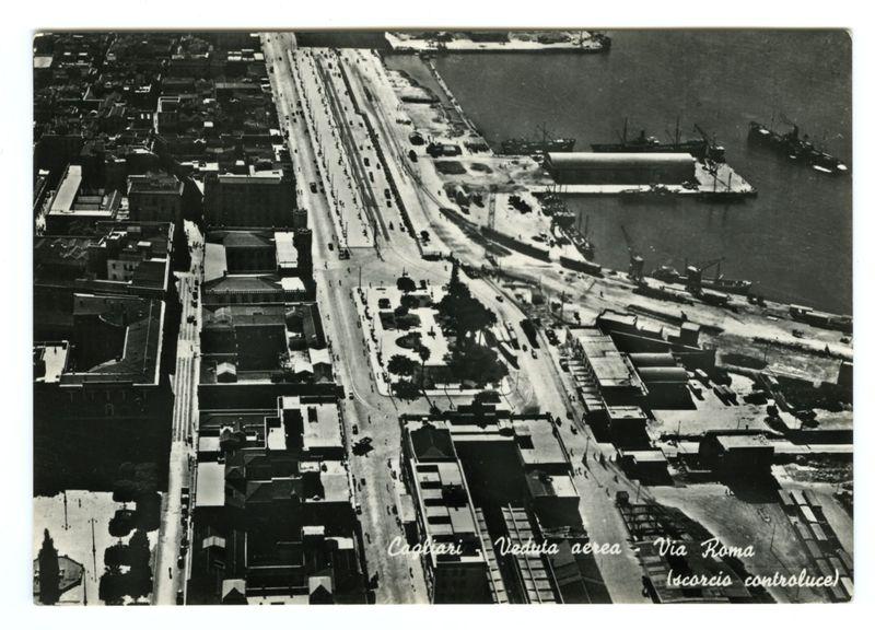 The image is a black and white aerial photograph depicting an urban landscape, likely taken during the early to mid-20th century given its monochrome nature. It shows dense city blocks with numerous buildings of various sizes, roads filled with vehicles indicating active traffic flow, and industrial areas such as docks or shipyards near what appears to be a waterfront.

The central road stretches from the foreground into the distance, flanked by rows of cars on both sides, suggesting it is an important thoroughfare. To its right are large open spaces that could serve as yards for storage or loading/unloading goods, possibly associated with maritime activities given their proximity to water and what looks like cranes in the background.

The photograph includes handwritten text overlaying parts of the image: "Cagliari Veduta aerea Via Roma (scorcio controluce)" which translates from Italian to English as "Aerial View Cagliari via Rome (darkened by smoke)." This indicates that the loc