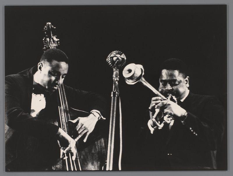The image is a black and white photograph showing two musicians in the midst of playing their instruments. On the left, there's an upright bass player wearing a dark suit with a bow tie; his fingers are positioned on the strings as he plays. His instrument has a large headstock at the top, typical for this type of jazz music ensemble. To his right is another musician playing what appears to be a trombone, judging by its shape and size, also dressed in formal attire. The background is mostly dark with no distinguishable features, focusing attention on the musicians themselves. Their intense expressions suggest they are deeply engaged in their performance.