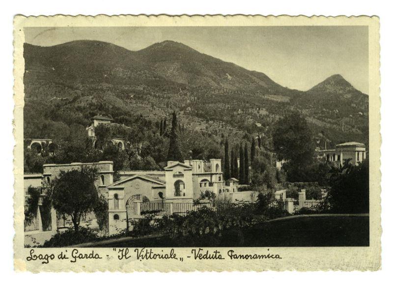 The image is a vintage black and white photograph of the landscape around "Il Vittoriale," located in Lago di Garda, specifically in Soagno di Garda. The scenery showcases rolling hills with dense vegetation and several distinct buildings that appear to be part of a grand estate or villa complex. Notable architectural features include archways, domed structures, and what seems like classical design elements on the façades of the houses.

The image has an aged appearance typical for vintage photographs, including slight discoloration at the edges where it's more worn out compared to the center. A handwritten caption in cursive script is present at the bottom left corner which reads "Soagno di Garda - 'Il Vittoriale,' - Veduta Panoramica," indicating that this photograph captures a panoramic view of Il Vittoriale.

The photo has an old-fashioned, historic feel to it, suggesting that it was taken in the early 20th century or late 19th century. It likely serves as either a historical r