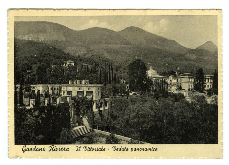 The image is a black and white vintage postcard depicting an old-fashioned panoramic view of the Vittoriale, located in Gardone Riviera. It features various architectural elements including classical buildings with ornate facades, arched windows, and decorative rooftops. The landscape includes rolling hills or low mountains partially covered by trees, which suggest a rural setting likely associated with historical significance.

The postcard has text at the bottom that reads "Gardone Riviera - Il Vittoriale - Veduta panoramica," indicating it's showcasing an extensive view of this location in Gardone Riveria. The overall aesthetic and style are indicative of early 20th-century photography or illustration, providing a glimpse into historical architecture and scenery from the region.

This image serves as visual representation for users who may not be able to see it directly but can infer details about its content through descriptions provided here: an old panoramic view postcard wit