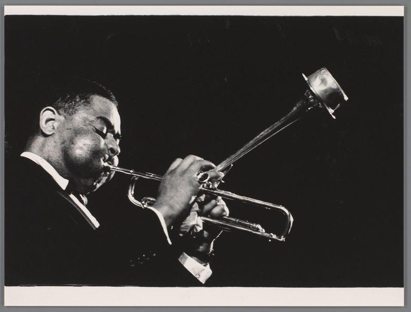 The image depicts a man playing the trumpet. He appears to be deeply focused on his performance, as evidenced by his eyes closed and mouth open in mid-blow. The photograph is monochromatic, rendered in black-and-white which highlights contrasts of light and shadow across his face and hands gripping the instrument. His attire suggests formality; he wears a suit jacket with notched lapels. This setting might be part of a musical performance or practice session given the spotlight effect on him against what appears to be a dark background, emphasizing his presence and action.

Additional context provided by the source indicates this photo captures Dizzy Gillespie during 'Jazz at the Philharmonic' in Scheveningen 1958. This historical reference situates it within jazz music's rich history, particularly highlighting one of its iconic figures.