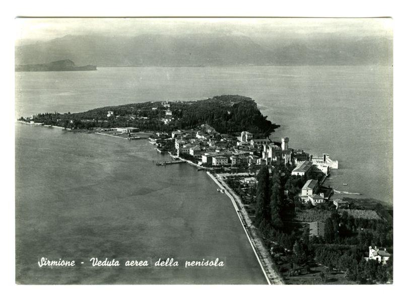 The image is an aerial photograph depicting the town of Sirmione on a peninsula extending into Lake Garda. The landscape features buildings, roads, and greenery with trees lining what appears to be paths or streets within the settlement. On one side of the peninsula, there's visible water that indicates it extends outwards from the lake. In the background, hills are faintly seen overcast by a grey sky suggesting an older photograph possibly taken in late summer or early fall given the absence of autumnal foliage colors and lack of snow on distant mountains.

There is text overlaid at the bottom left corner with "Sirmione - Veduta aerea della penisola," which translates to Sirmione - aerial view of the peninsula. This suggests that the photograph might be historical, showing an older or preserved state of this lakeside town and its surroundings before significant modern development occurred.

The black-and-white nature of the image indicates it is not recent but rather from several 