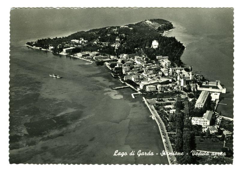 This black and white aerial photograph depicts an expansive view of the Sirmione peninsula, situated on Lake Garda in Italy. The town is densely packed with buildings exhibiting a variety of architectural styles ranging from traditional European designs to neoclassical structures. A notable feature includes a large castle-like edifice at one end, suggesting historical significance or importance.

The waterfront shows several piers and docks extending into the lake, indicating active use for boating activities. There's also a small boat with two occupants near the shore on the left side of the photo, likely engaging in leisurely water-based recreation.

Greenery is abundant along the coastline, providing a lush contrast to the urban landscape, indicative of well-maintained parks or gardens which seem vital for relaxation and perhaps as an escape from the town's built environment. The absence of modern vehicles suggests this image could be several decades old, capturing life in Sirmi