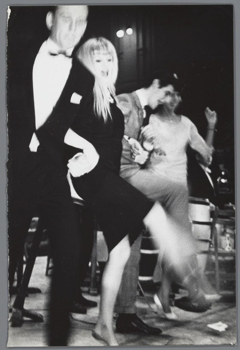 The image depicts a lively dance scene, captured in black and white. In the foreground, there is a woman with short hair wearing a dark dress dancing energetically on her toes; she appears to be mid-twirl or leap, with one leg extended back behind her and arms outstretched for balance. Her facial expression suggests excitement or joy.

Behind her, another person in casual attire seems caught off-balance, possibly reacting to the woman's movement. This individual is wearing a light-colored top and dark pants, indicating an informal setting. The background includes chairs that hint at seating arrangements typical of dance venues like nightclubs or ballrooms.

The image has a candid quality with motion blur on some subjects, emphasizing the spontaneity of the moment captured. There are blurred shapes in the far distance which could be other dancers or people seated away from the action. No discernible text is present to provide context about the location or event taking place.

This