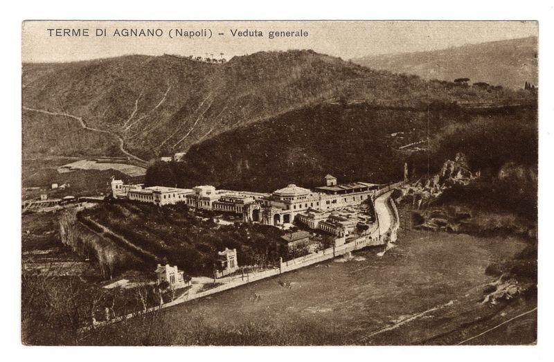 The image is a sepia-toned photograph depicting an aerial view of Terme Di Agnano in Naples, labeled as "Veduta generale." The scene captures the historical site with its sprawling complex nestled within hilly terrain. Several large buildings and structures are visible, characterized by classical architecture, including domes and columns, likely representing thermal baths or spa facilities typical of such sites during that era. A winding road snakes through the landscape, leading up to a hilltop where more buildings can be seen in silhouette against the sky. The vegetation around is sparse, with trees dotting some areas on either side of the structures. There's no visible human activity within this view; it seems like an early 20th-century snapshot providing historical context without any modern-day interruptions or distortions.
