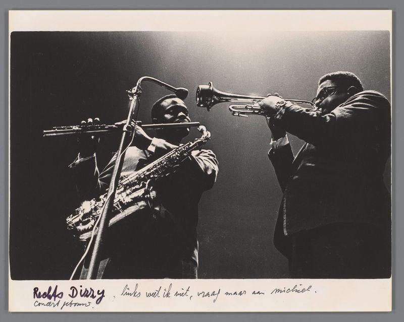 The image is a black and white photograph capturing two musicians in the midst of a performance. The musician on the left plays an upright bass, while the one on the right holds onto his trumpet with both hands as if he's either playing or has just finished playing it. They are standing against what appears to be a dark background that could represent smoke from their instruments. Below them is some floral decoration, perhaps part of the stage setting for a concert.

The photograph includes handwritten text in Dutch at the bottom which translates roughly to "Dizzy's Diary," with additional notes about improvisation and mention of Michel. The handwriting suggests it might be an autograph or inscription by someone named Ed van der Elsken who is known as a photographer, possibly indicating that this image was taken during one of his photography sessions.

The photograph has been annotated at the top left corner which says "Redds Diary - links wet ik met, vroly maar aan michael," likel
