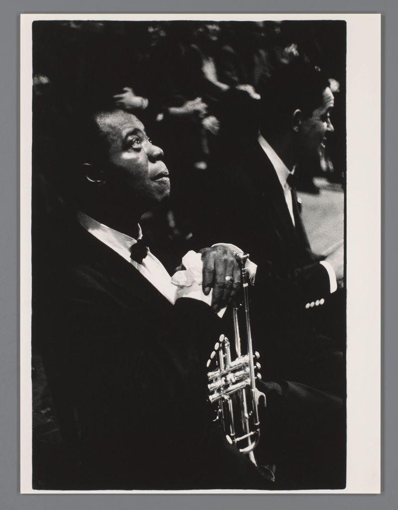 The image is a black and white photograph featuring Louis Armstrong. He appears seated, holding his iconic trumpet in one hand while the other rests on what looks to be a music stand or similar object. His expression seems contemplative as he gazes off to the side rather than directly at the camera.

In terms of visual elements:
- The main subject is centered and occupies most of the frame.
- Louis Armstrong wears formal attire, including a dark suit with a bow tie.
- A second figure in partial view appears behind him, out of focus, suggesting an audience or another musician.
- The trumpet's reflective surface catches light, creating bright highlights on its valves and tubing.

Technical details:
- Image quality is grainy indicative of the film stock used at that time (e.g., Ilford HP5).
- Composition focuses sharply on Armstrong with a blurred background enhancing his prominence in the scene.