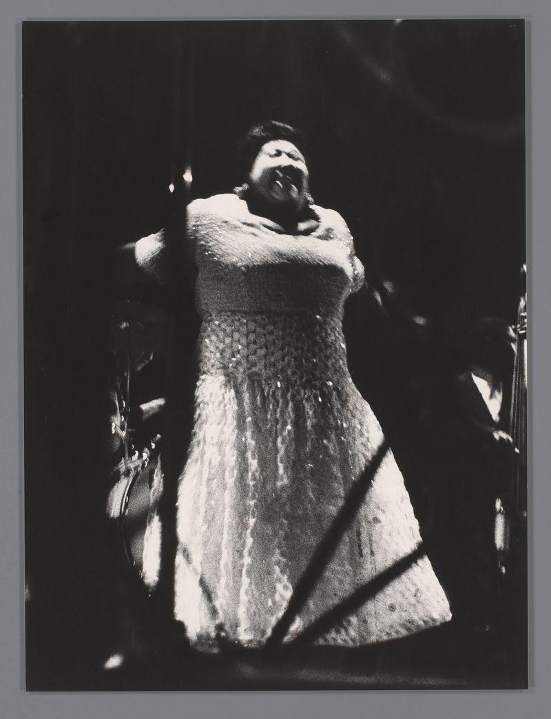 The image is a black and white photograph capturing an individual in the midst of singing. The person, who appears to be female based on attire and physical features, is dressed in what seems like a light-colored dress with ruffled details at the waistline. Her arms are raised above her head as she holds onto them or possibly another performer beside her, emphasizing the intensity of the performance moment captured.

The image's composition focuses on the individual against a dark background that lacks any discernible elements apart from shadows and highlights created by stage lighting. The play of light falls across different parts of her dress and body, accentuating textures such as ruffles or folds in the fabric.

There is no explicit context provided about the location beyond suggesting it could be an indoor performance space due to the dark backdrop which typically implies a concert hall setting. Notably, there's a watermark at the top right corner that indicates copyright by 