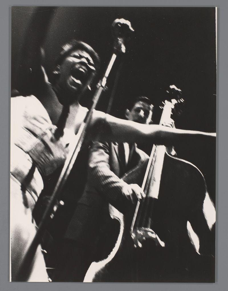 The image depicts a monochrome photograph of two musicians performing, with the foreground artist captured in an expressive pose, seemingly singing into a microphone. The musician's face is contorted in what appears to be an intense vocal performance, mouth wide open as if capturing a note or emotion powerfully. They are dressed casually yet performantly, wearing a suit jacket and a loose-fitting shirt that suggests movement during the act.

The background artist is partially visible with their focus directed towards playing a double bass, captured in mid-stroke which indicates active engagement in the musical piece being performed. The lighting accentuates the dramatic expressions on both faces while casting deep shadows around them, adding to the emotive quality of the image. There's an evident energy conveyed through their gestures and body language.

The background is dark, drawing attention to the figures and enhancing the contrast between light and shadow in this dynamic scen